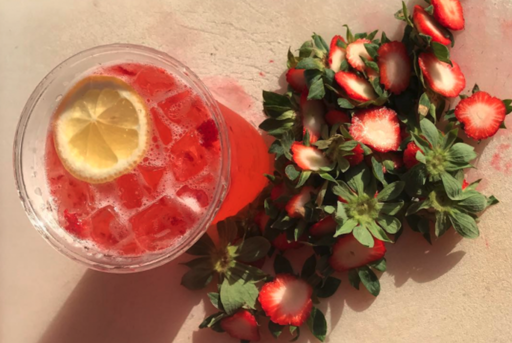Strawberry lemonade in a container with a lemon slice and ice, chopped up strawberries lay in an adjacent pile