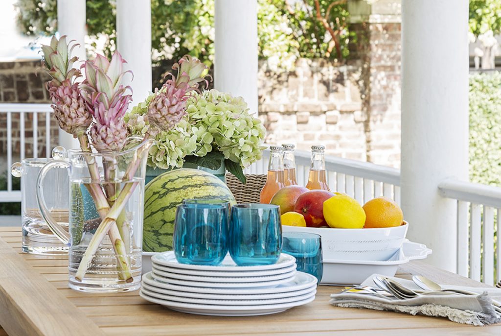 Beautiful outdoor dining setting with blue glasses, white plates, florals, a fruit bowl, a watermelon, utensils, and some bottled drinks set on the table