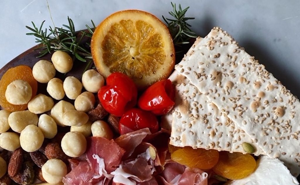 Crackers, nuts, peppers, herbs, and meat are seen on a charcuterie board